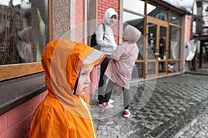 Portrait cute little lonely caucasian kid boy in bright orange waterproof raincoat stand under rain against brick wall