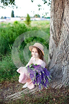 Portrait of a cute little smiling brunette with long hair sitting near a tree with a huge bouquet of purple lupins on a golden sun