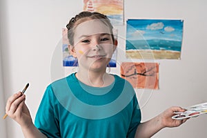 Portrait of cute little teenage girl with brush and palette on background of her drawings. A young artist with a face