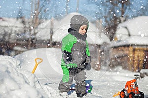 Portrait of cute little toddler sitting on snow and playing with his yellow tractor toy in the park. Child playing