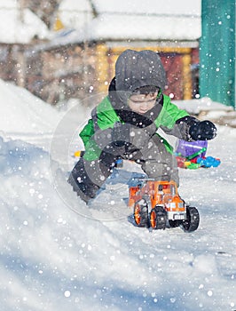Portrait of cute little toddler sitting on snow and playing with his yellow tractor toy in the park. Child playing