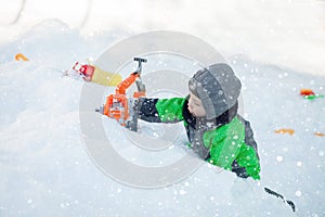 Portrait of cute little toddler sitting on snow and playing with his yellow tractor toy in the park. Child playing outdoors. Happy