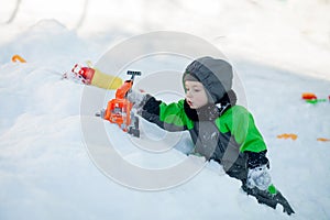 Portrait of cute little toddler sitting on snow and playing with his yellow tractor toy in the park. Child playing outdoors. Happy