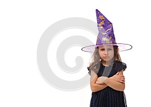 Portrait of cute little witch angry upset girl wearing a wizard hat and dressed in stylish carnival dress, looking at camera