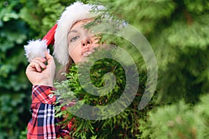 Portrait of cute lovely gir in a santa claus red hat sending blowing kiss with pout lips looking at camera through green branches.