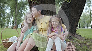 Portrait cute mature woman kissing her two cute granddaughters sitting on the grass under the tree in the park, looking