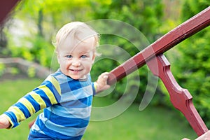 Portrait of cute mischievous caucasian blond baby boy holding wooden banister climbing staircase at outdoor backyard playground.