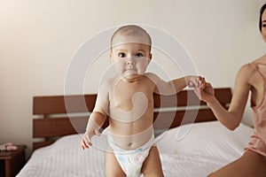 Portrait of cute newborn baby standing on bed in morning mother holding her child hand.