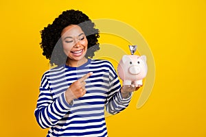 Portrait of cute optimistic girl with chevelure wear striped pullover look directing at money box isolated on yellow