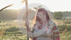 Portrait cute overweight woman holding a scythe looking at camera on the green summer field in sunlight. Beautiful