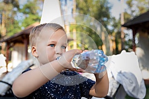 Portrait of cute playful little caucasian blond thirsty toddler boy kid holding plastic bottle and drinking water on hot