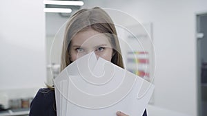 Portrait of cute playful young lady in formal clothes hiding her face behind papers looking in camera and away. Woman