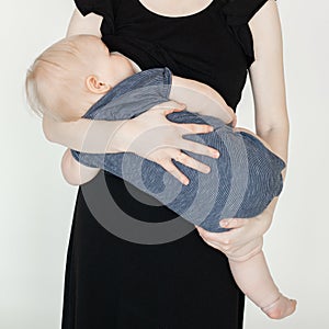 Portrait of cute plump cherubic baby infant toddler in grey bodysuit holded by mother in black dress eating breast milk.