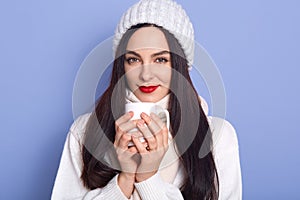 Portrait of cute positive brunette woman wearing white warm cap and sweater, female holding cup of coffee or tea, posing isolated