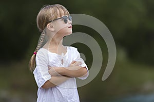 Portrait of cute pretty funny young girl with blond braids in white dress and dark sunglasses.