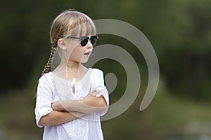 Portrait of cute pretty funny young girl with blond braids in white dress and dark sunglasses.