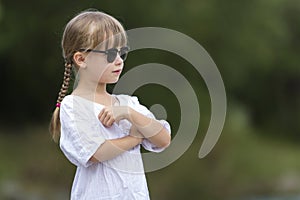 Portrait of cute pretty funny young girl with blond braids in white dress and dark sunglasses