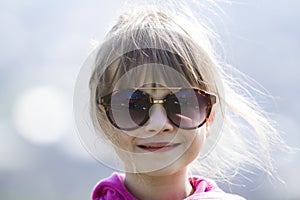 Portrait of cute pretty little blond preschool girl in pink sweater and dark sunglasses smiling happily in camera with funny