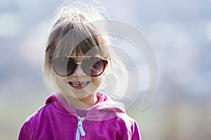 Portrait of cute pretty little blond preschool girl in pink sweater and dark sunglasses smiling happily in camera with funny child