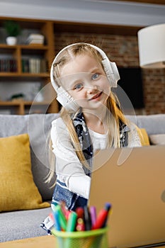 Portrait of cute primary school girl studying at home using laptop computer. Smiling child wearing white earphones