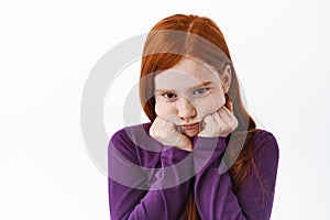 Portrait of cute redhead girl pouting, making coy sulking face and look gloomy from under forehead, standing upset