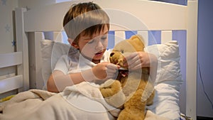 Portrait of cute sick boy measuring temperature to his toy teddy bear with digital thermometer. Concept of child virus