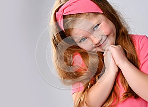 Portrait of cute, sly smiling redhead kid girl with freckles on face in pink t-shirt and headband holding hands together