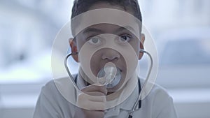 Portrait of cute small boy with stethoscope in his ears looking in camera while breathing in device. Healthcare, healthy