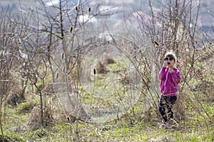 Portrait of cute small confused blond girl in casual pink clothing and dark sunglasses standing alone lost among dry prickly bush