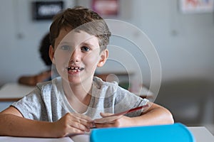 Portrait of cute smiling caucasian elementary schoolboy with pencil sitting at desk in classroom