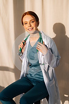 Portrait of cute smiling female doctor in white coat sitting on chair near window in sunny day in medical clinic office.