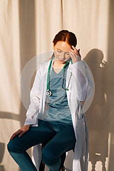 Portrait of cute smiling female doctor in white coat sitting on chair near window in sunny day in medical clinic office.