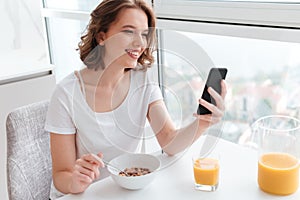 Portrait of cute smiling girl in white tshirt chatting on smartphone while sitting and eating cornflakes at the kitchen table