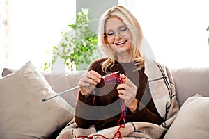 Portrait of cute smiling grandmother with blonde hair wearing comfy pullover when knitting wool handmade socks sitting