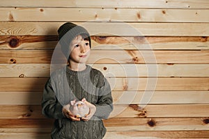 Portrait of cute smiling little boy with piggy bank on wooden background. Child with money in green hat.