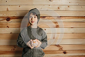 Portrait of cute smiling little boy with piggy bank on wooden background. Child with money in green hat.