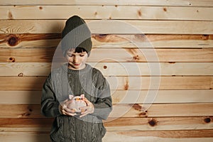 Portrait of cute smiling little boy with piggy bank on wooden background. Child with money in green hat.