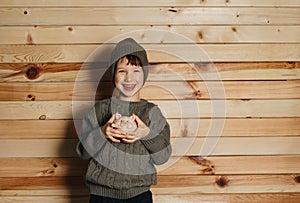 Portrait of cute smiling little boy with piggy bank on wooden background. Child with money in green hat.