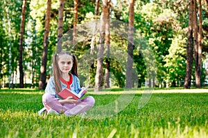 Portrait of Cute Smiling Little Girl Sitting On Grass And Reading Book In Park