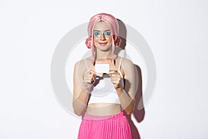 Portrait of cute smiling party girl in pink wig, showing credit card, standing over white background