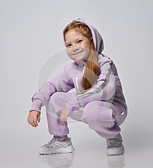 Portrait of cute smiling red-haired kid girl in pink jumpsuit sitting squatted with her hood on her head