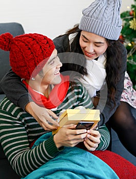 Portrait of cute smiling young Asian lover couple in long sleeve sweatshirt, scarf, knitted hat sitting on a floor near Christmas