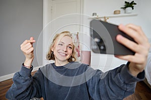 Portrait of cute smiling young woman with smartphone, taking selfies in her room, showing heart sign, taking pictures or