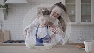 Portrait cute son helps mom in the kitchen. The boy interferes with the milk in a glass bowl using whisks.