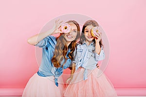 Portrait of cute surprised young woman and little long-haired girl having fun with donuts on pink background. Stylish