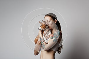 Portrait of a cute tattooed young woman hugging and kissing her little puppy basenji dog. Love between dog and owner