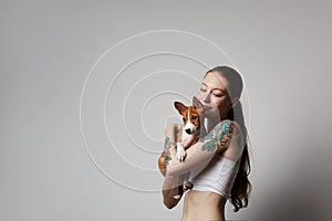 Portrait of a cute tattooed young woman hugging and kissing her little puppy basenji dog. Love between dog and owner