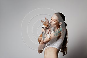 Portrait of a cute tattooed young woman hugging and kissing her little puppy basenji dog. Love between dog and owner