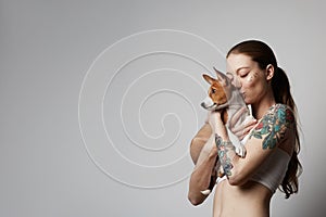 Portrait of a cute tattooed young woman hugging and kissing her little puppy basenji dog. Love between dog and owner