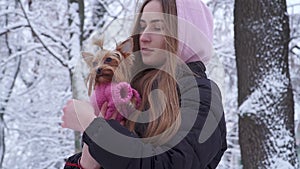 Portrait cute teen girl with long hair covered with a hood hugging a yorkshire terrier dressed in wool sweater holding